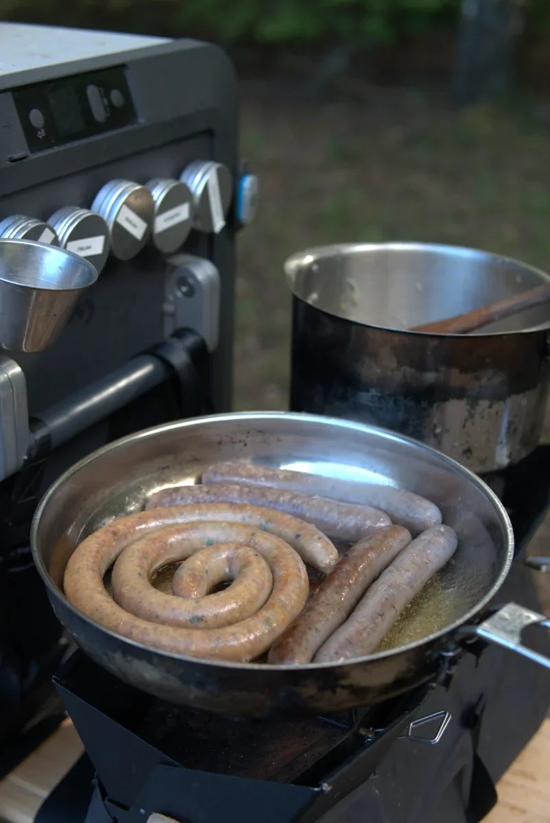 risotto puttrar på medan korv steks i stekpanna på campingkök med kylbox i bakgrunden